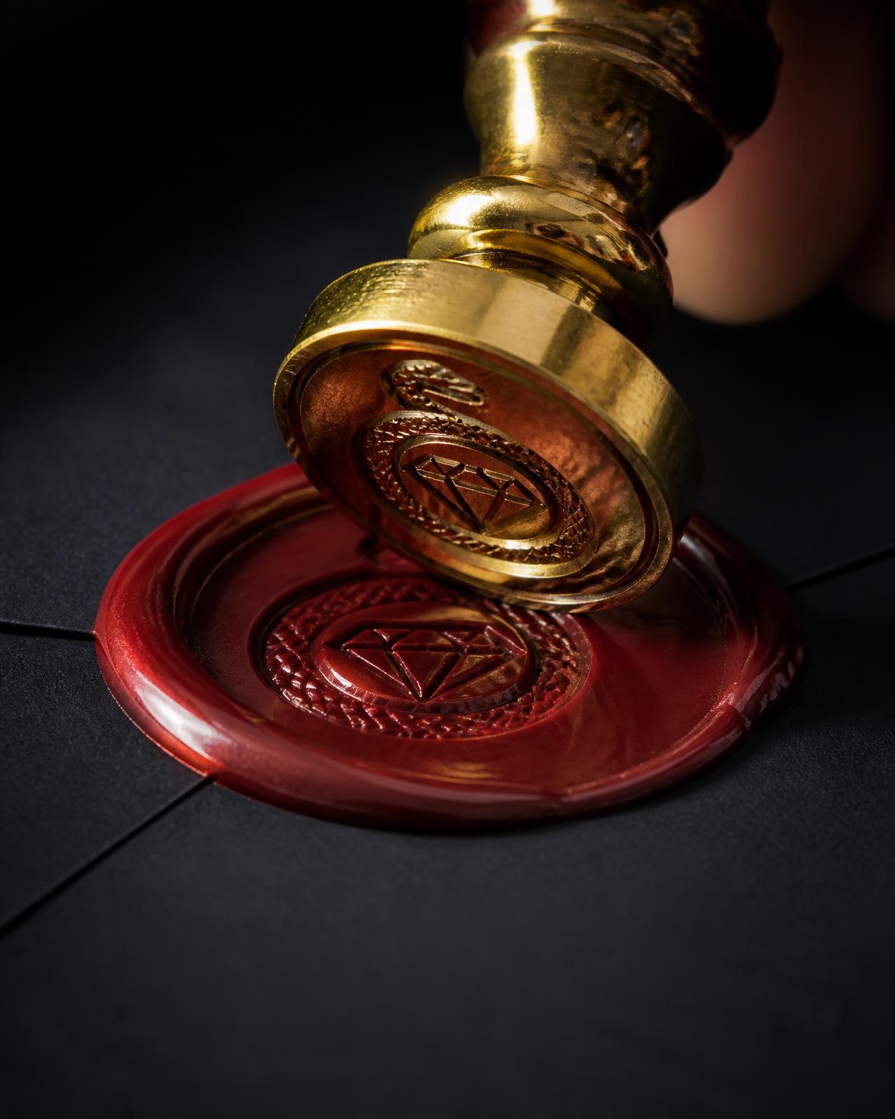 Brass serpent seal pressing an ornate crest into deep red wax on black paper
